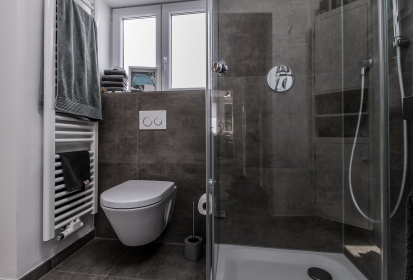 Grau gefliestes Bad mit Eckdusche und daneben Toilette unter Fenster.jpg