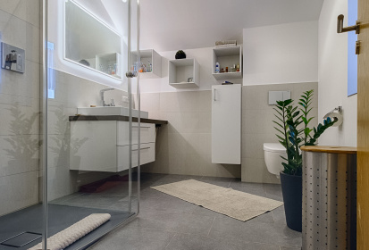 Badezimmer mit grauen Fliesen, graue Duschtasse, Waschbecken mit Unterschrank, eckige wuerfelfoermige Badezimmermoebel, versteckte Toilette.jpg