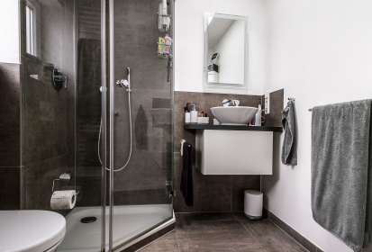 Ein dunkelgrau gefliestes Bad mit Dusche in der Ecke und schmalem, elegantem Waschbecken daneben.jpg