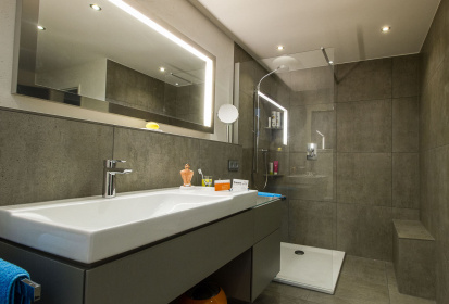 Ein Traum in grau – Blick auf das weisse Waschbecken mit grauem, buendigen Unterschrank und dahinter die quadratische ebenerdige Dusche mit weisser Duschtasse und Sitzmoeglichkeit.jpg